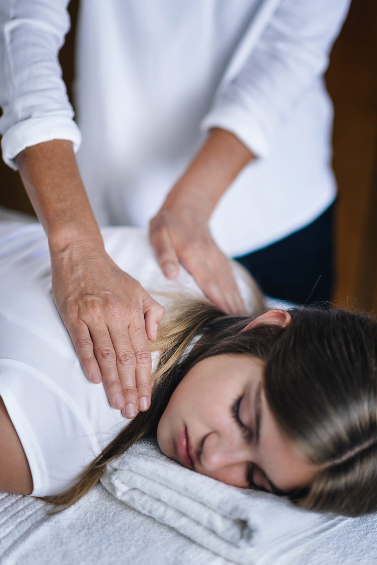 Vertical image of female Reiki therapist holding hands over shoulders of the patient and transfer energy. Peaceful teenage girl lying with her eyes closed. Alternative therapy concept of stress reduction and relaxation.